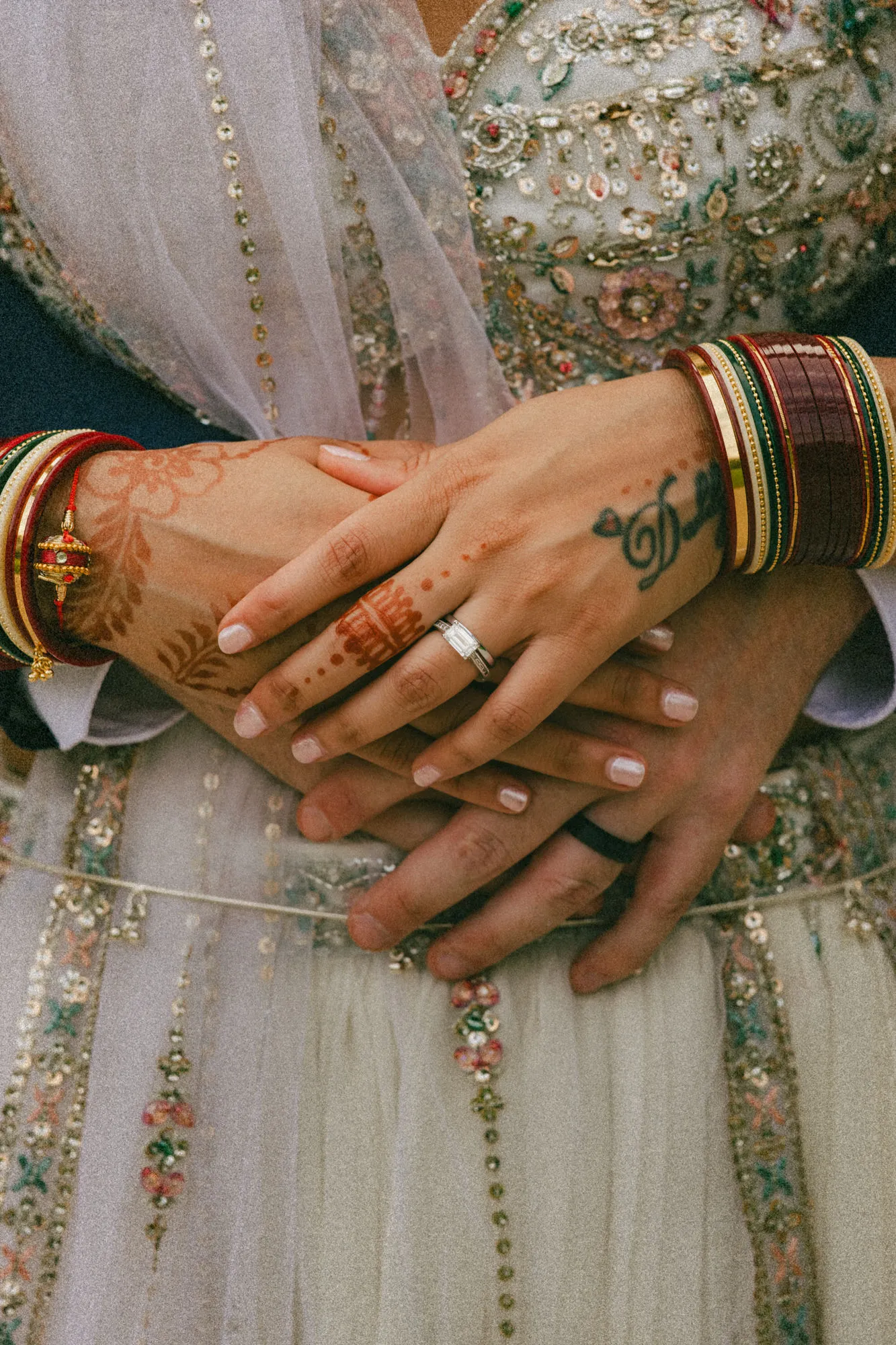 Grainy macro of stacked hands with henna, red chura bangles and both wedding rings, by AD Photography