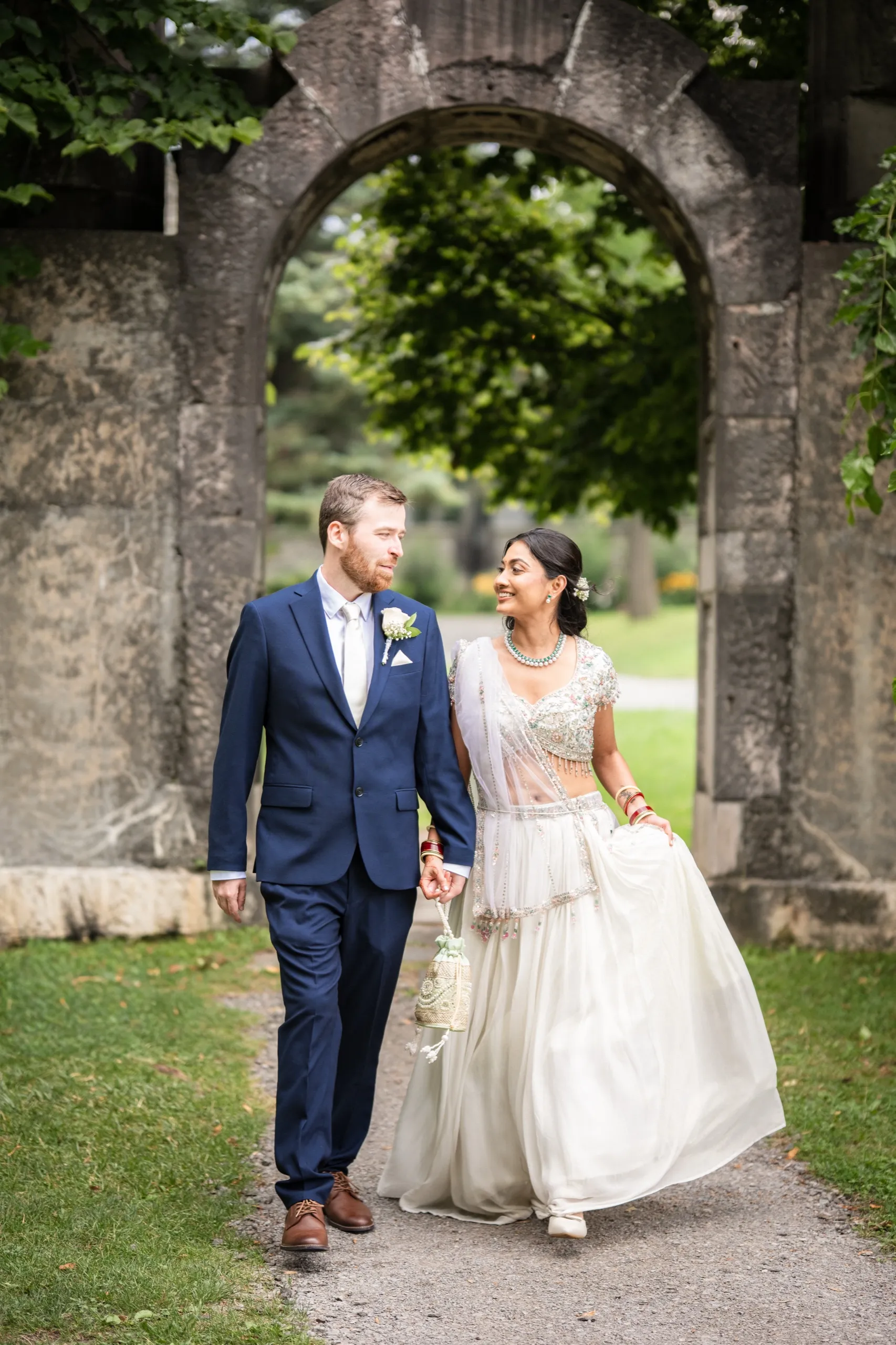 Nidhi and Derek walk hand in hand through a stone archway, trading a smile., by AD Photography