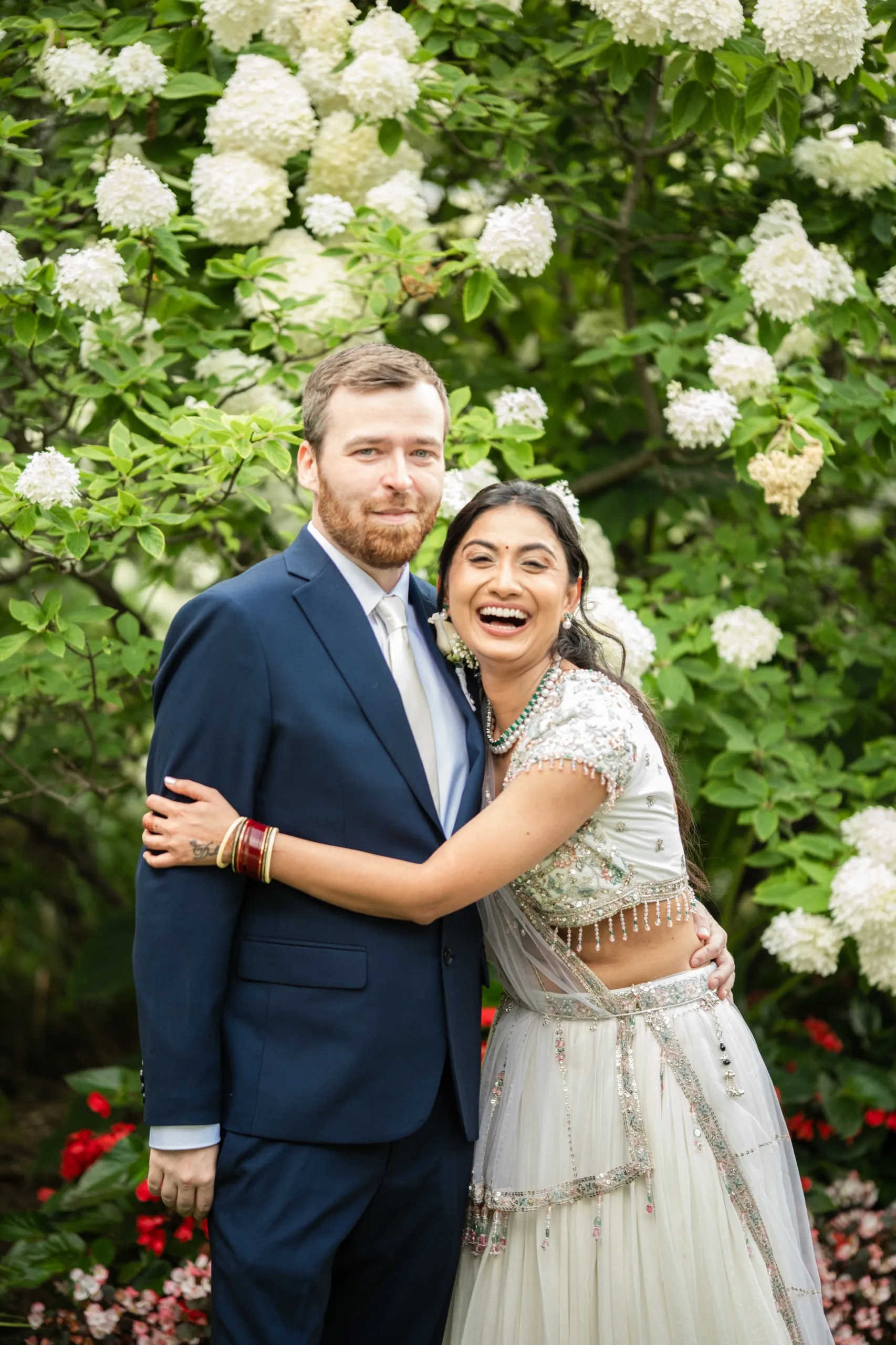 Nidhi laughs freely as Derek holds her close beneath a cloud of white hydrangea., by AD Photography