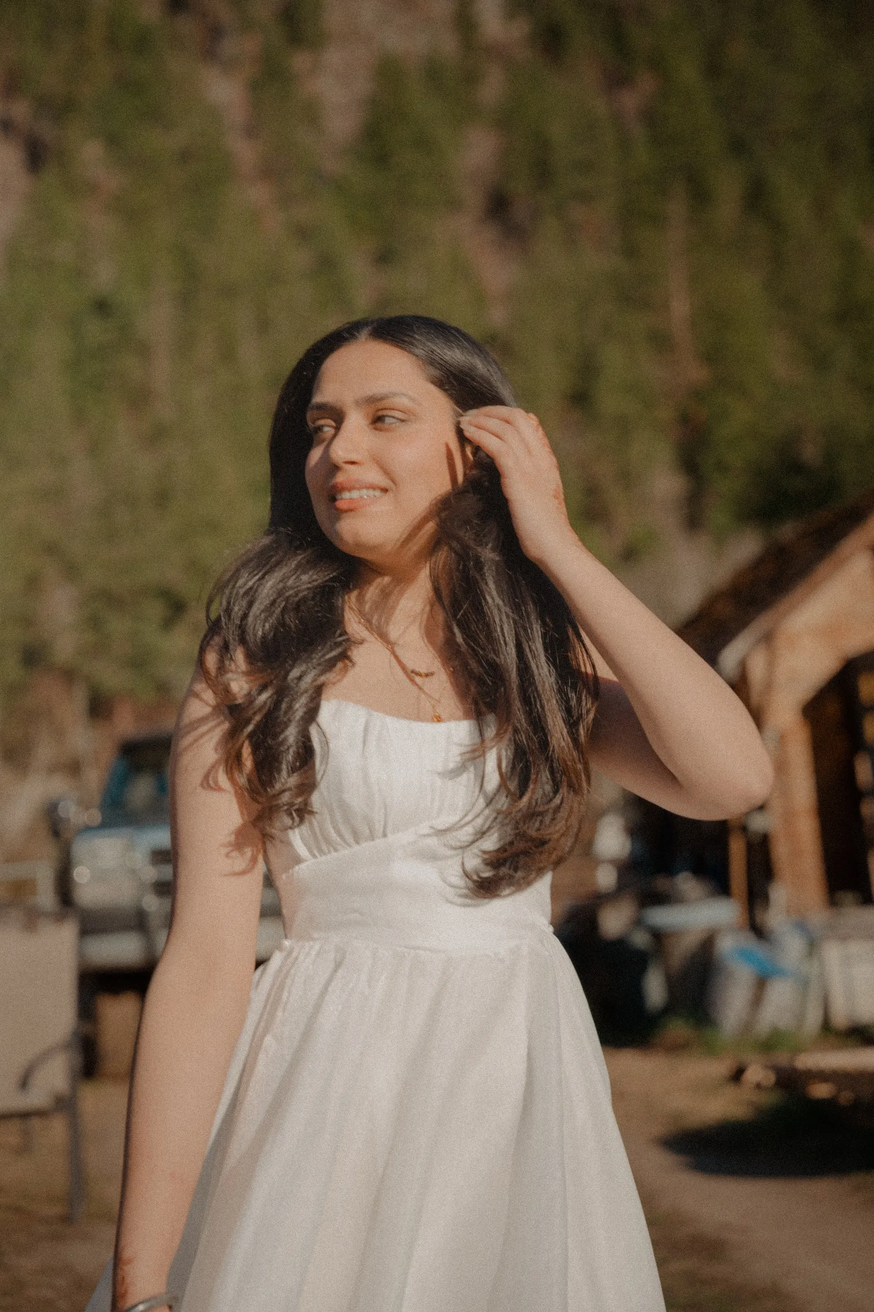 Mehak tucking her hair back in a white sundress outside an old log cabin, by AD Photography