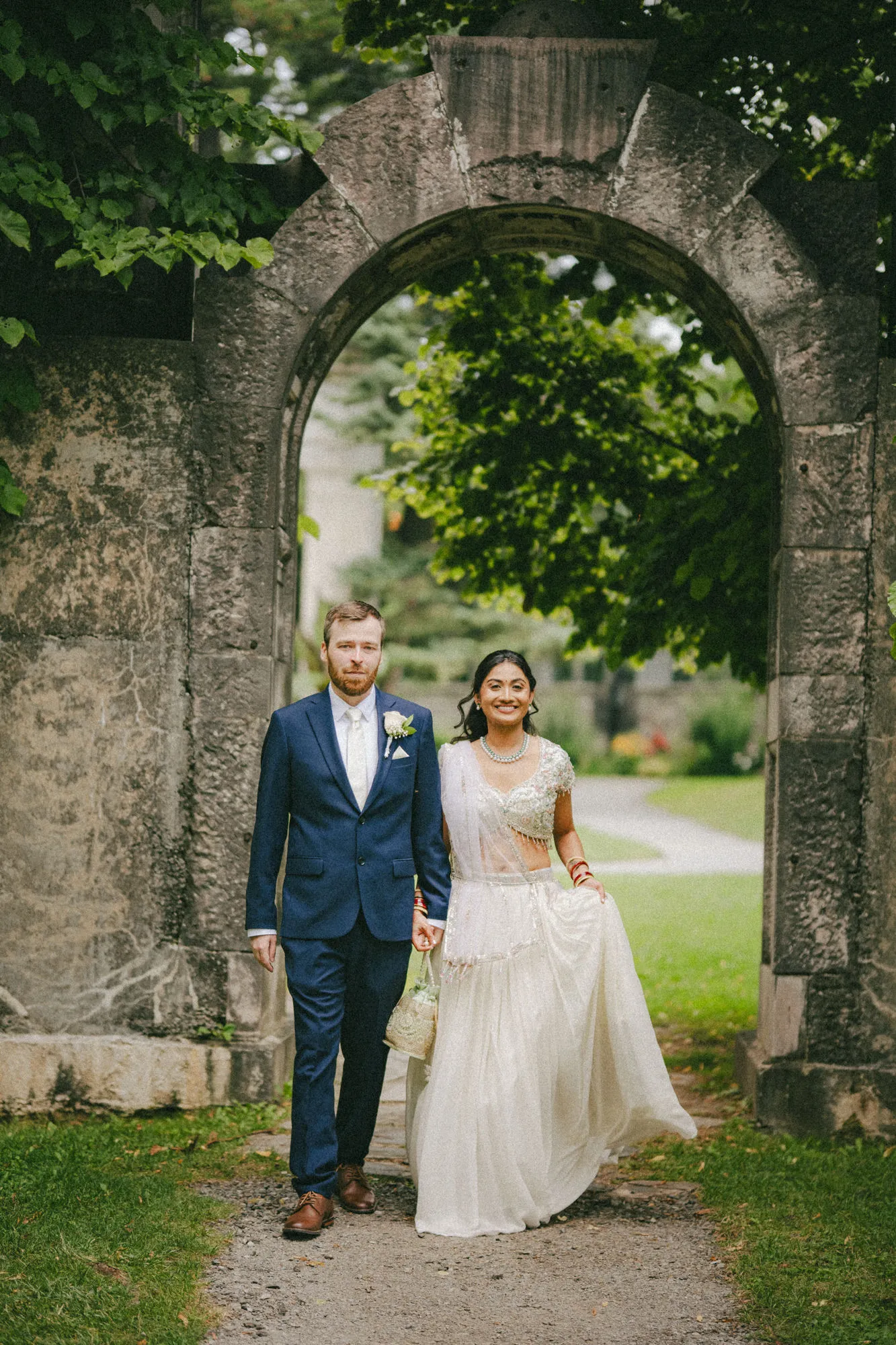 Nidhi and Derek pause hand in hand beneath the salvaged stone gateway arch, by AD Photography