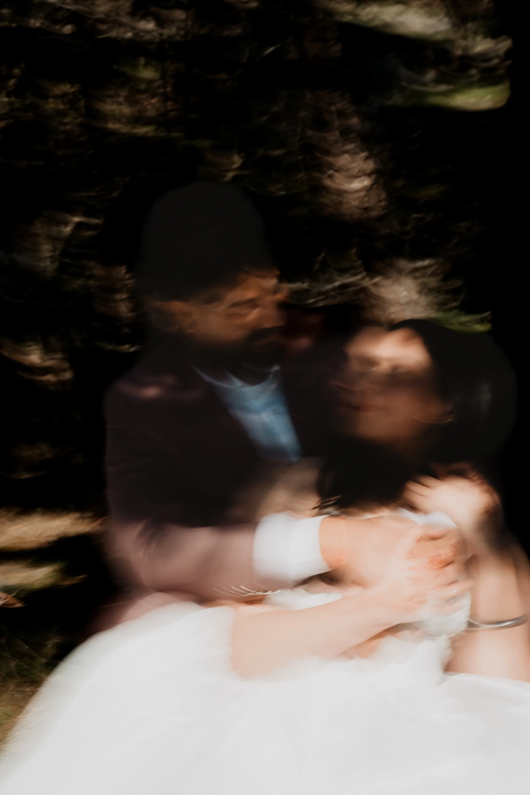 Motion blur of the couple wrapped together in deep shade beneath the trees, by AD Photography
