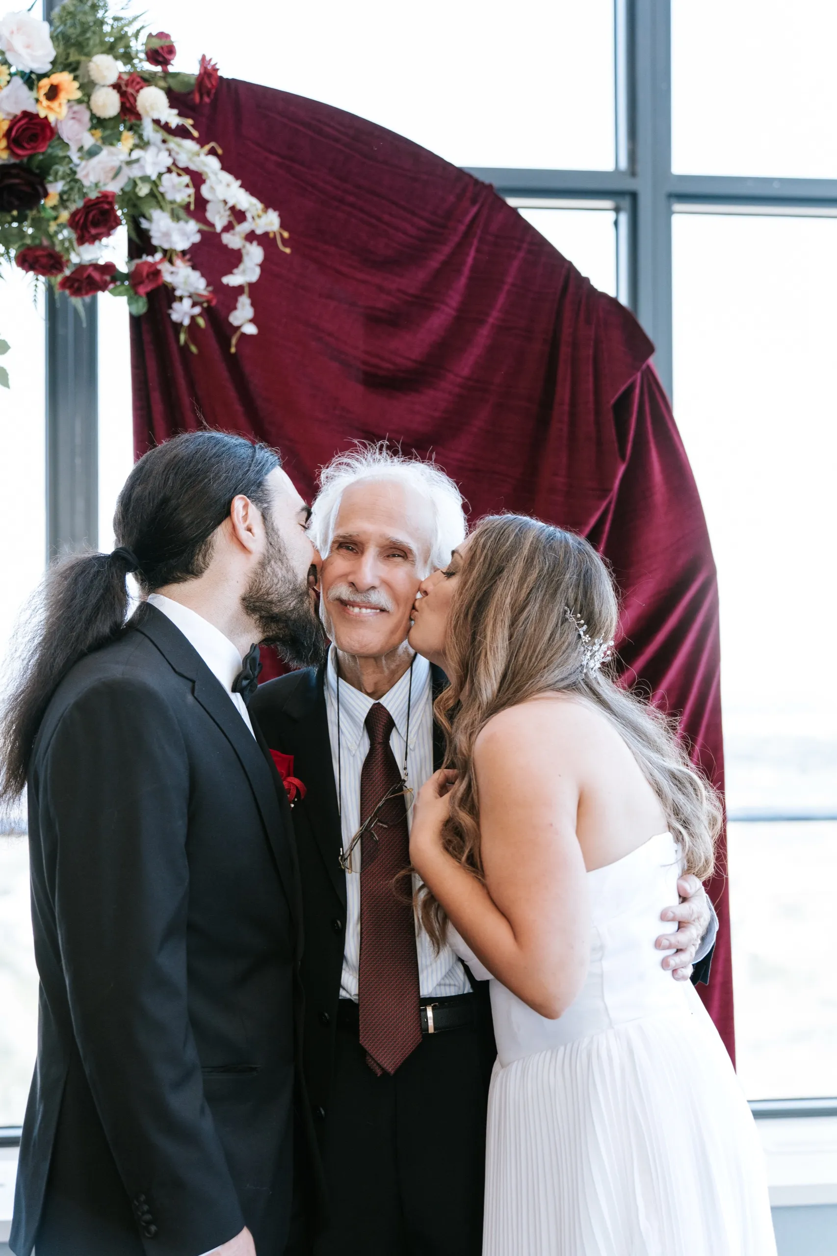 Zoha and Moe kiss a family elder on both cheeks in front of the burgundy drape, by AD Photography