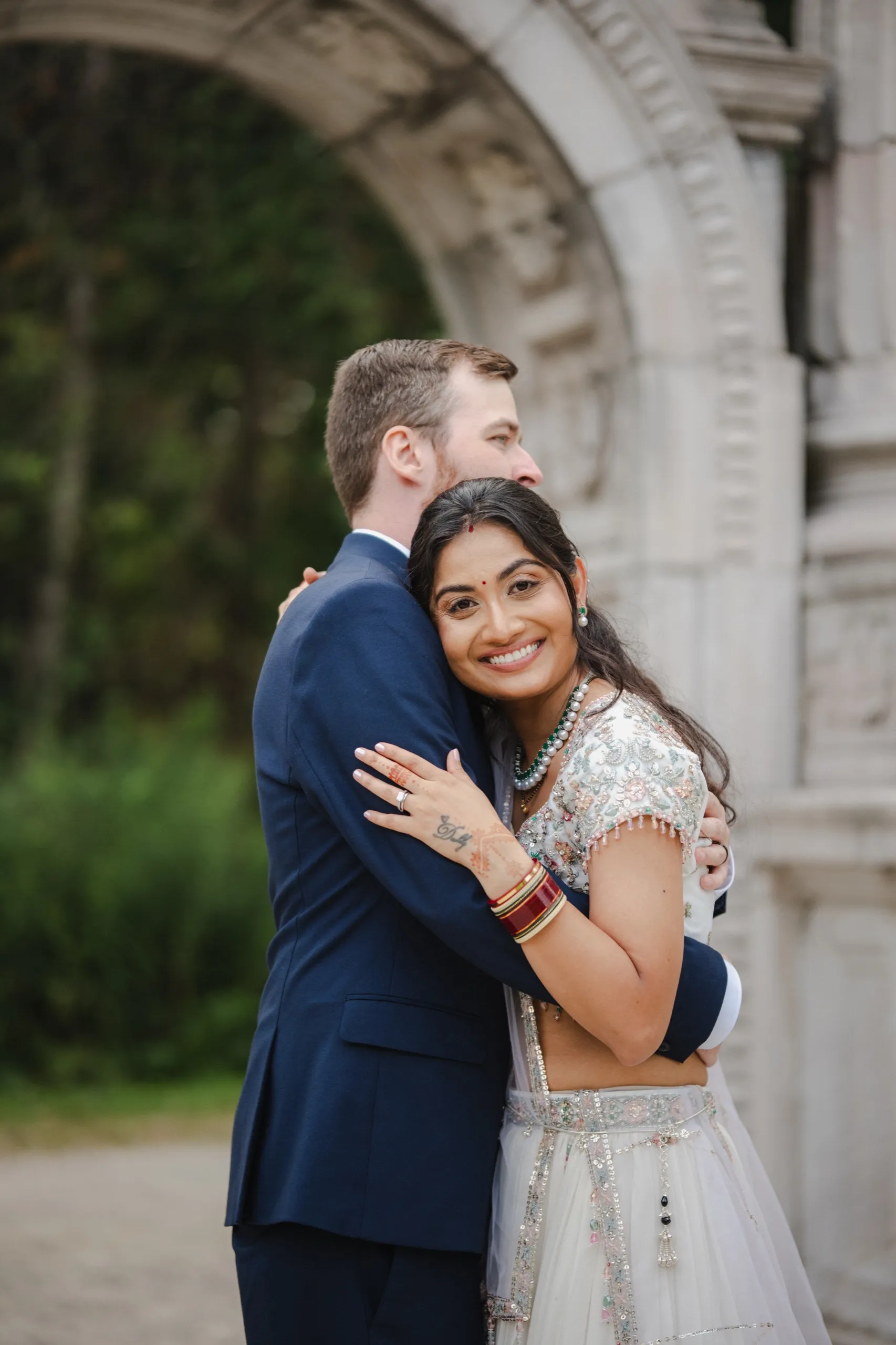 Nidhi hugs Derek close and grins at the camera in front of the stone arch., by AD Photography