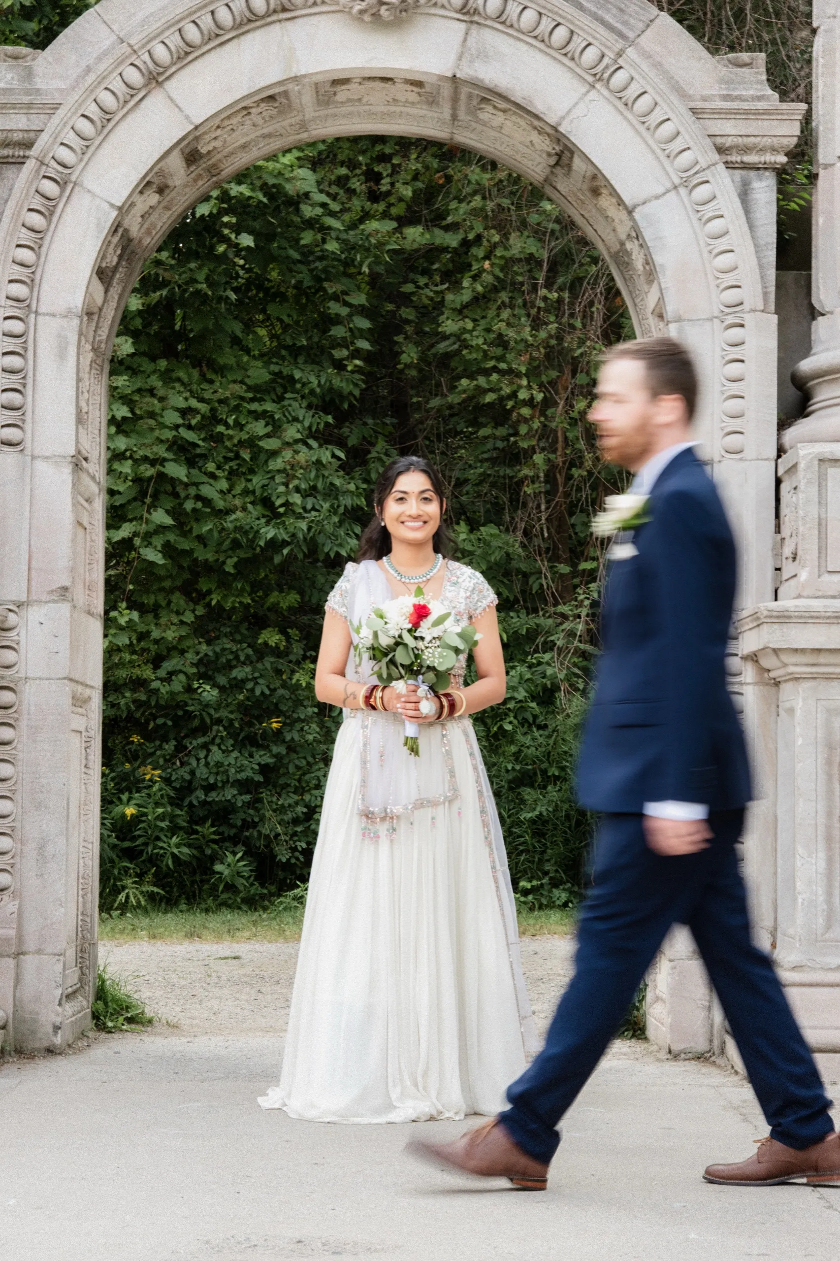 Grainy motion blur of Derek walking past as Nidhi waits smiling beneath the arch., by AD Photography