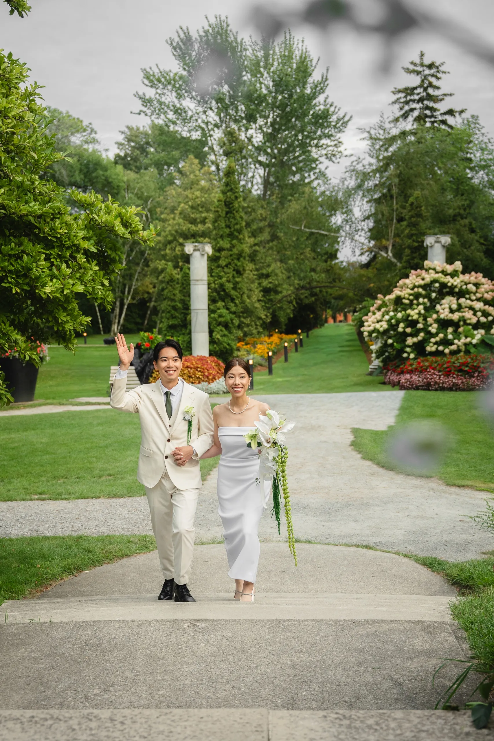 Shot through foreground leaves as the couple walk the columned garden allee, Nelson waving, by AD Photography