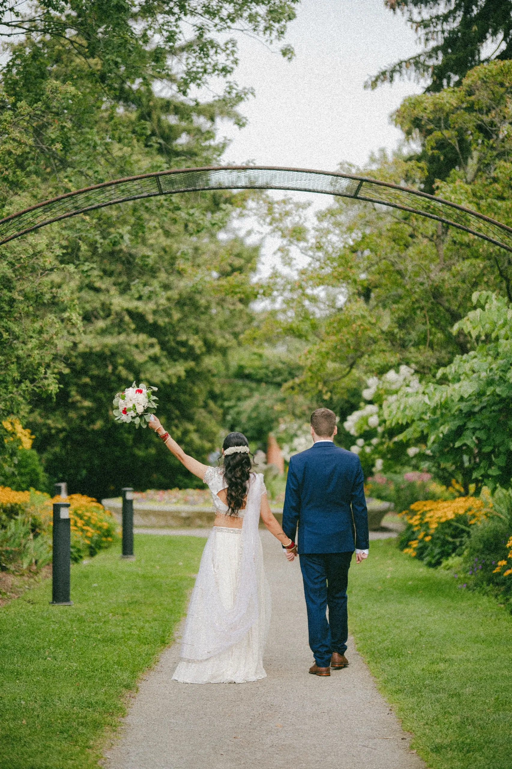 Nidhi raises her bouquet high as she and Derek walk beneath a garden arch., by AD Photography