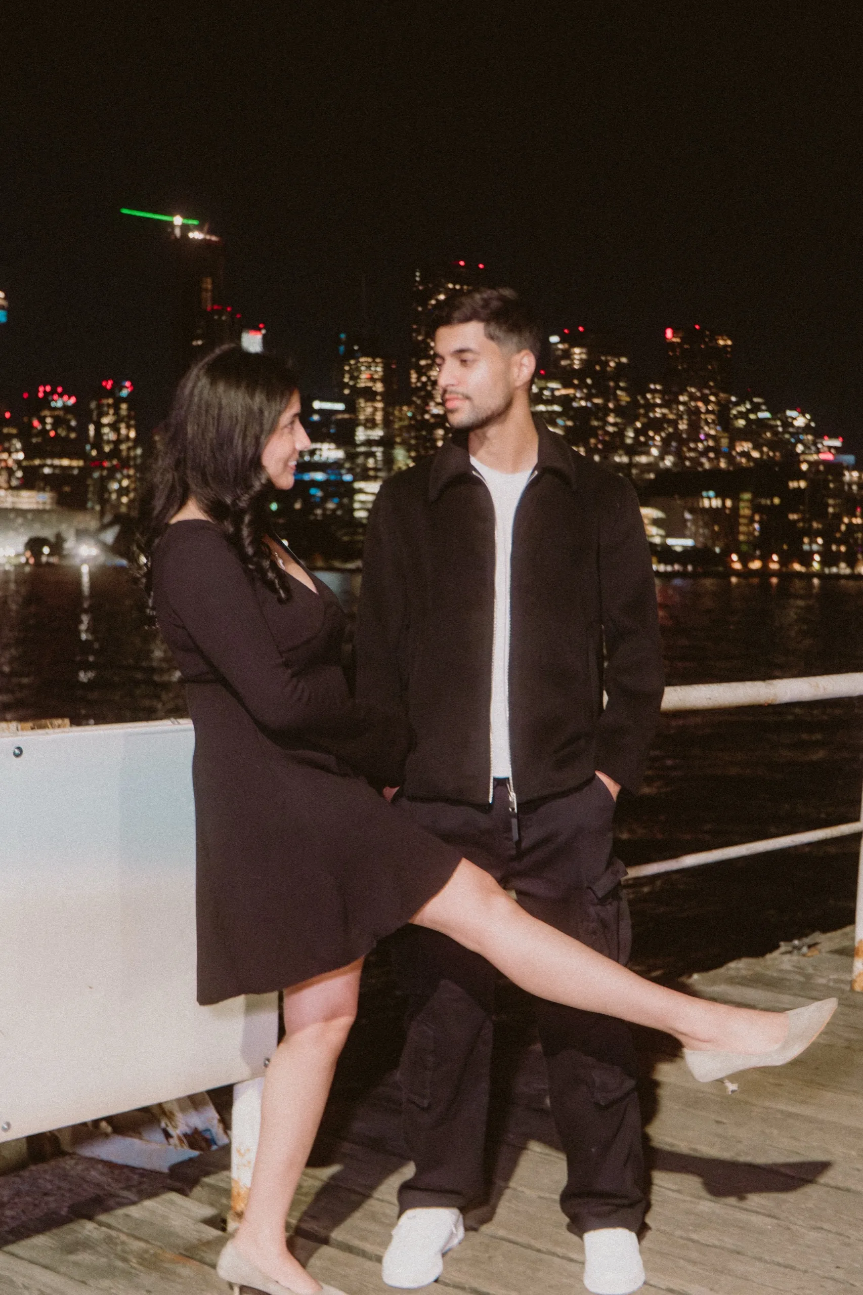 Swati swings a playful leg across Saksham on the pier, city lights behind, by AD Photography
