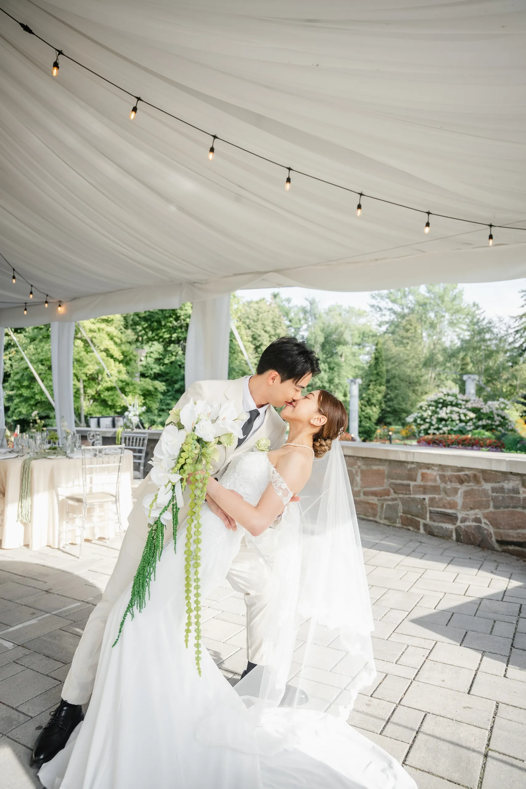A dip and a kiss under the string lit wedding reception tent, bouquet cascading green, by AD Photography