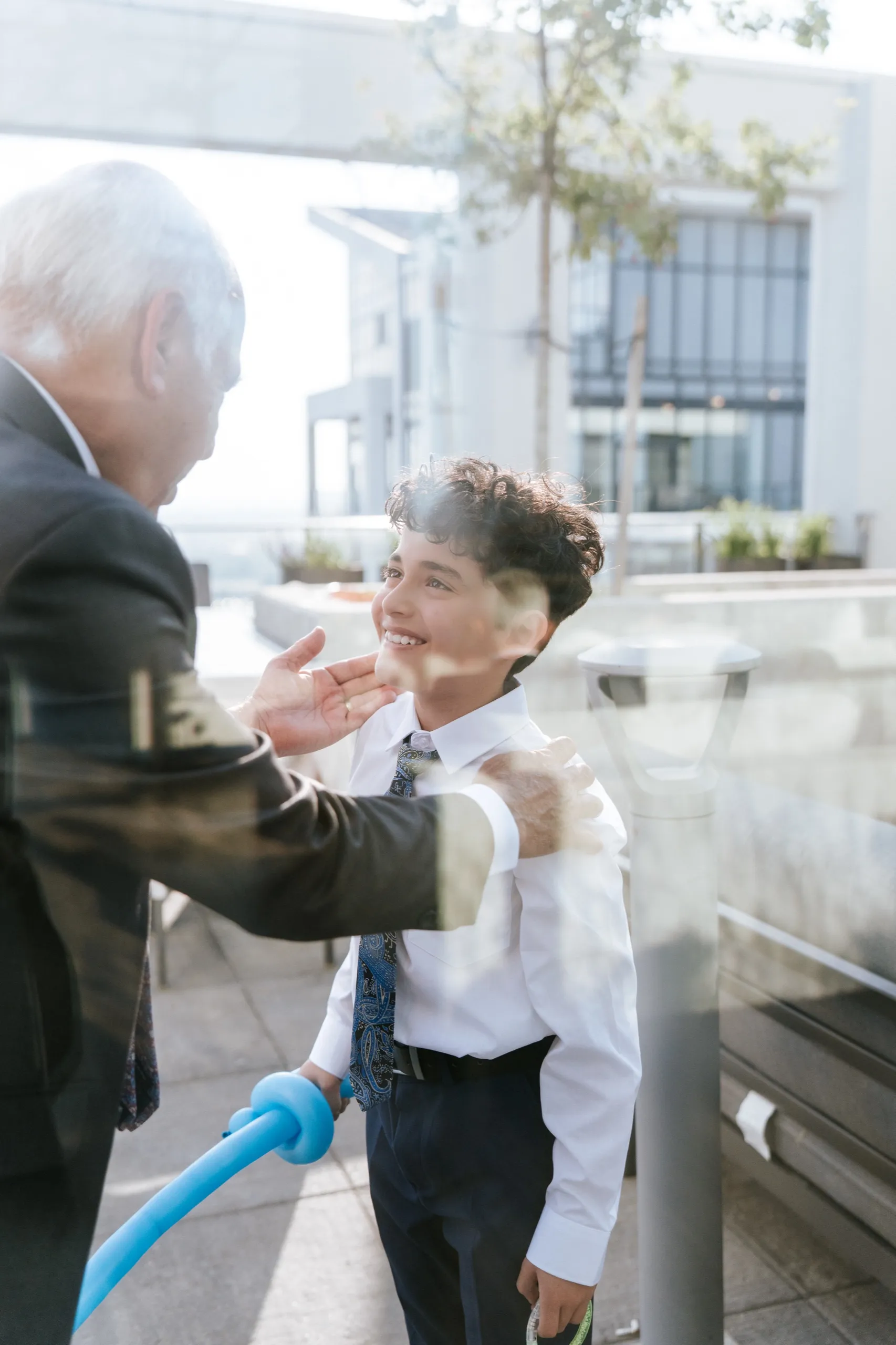 An elder pinches a boy's cheek out on the terrace, caught through the glass, by AD Photography