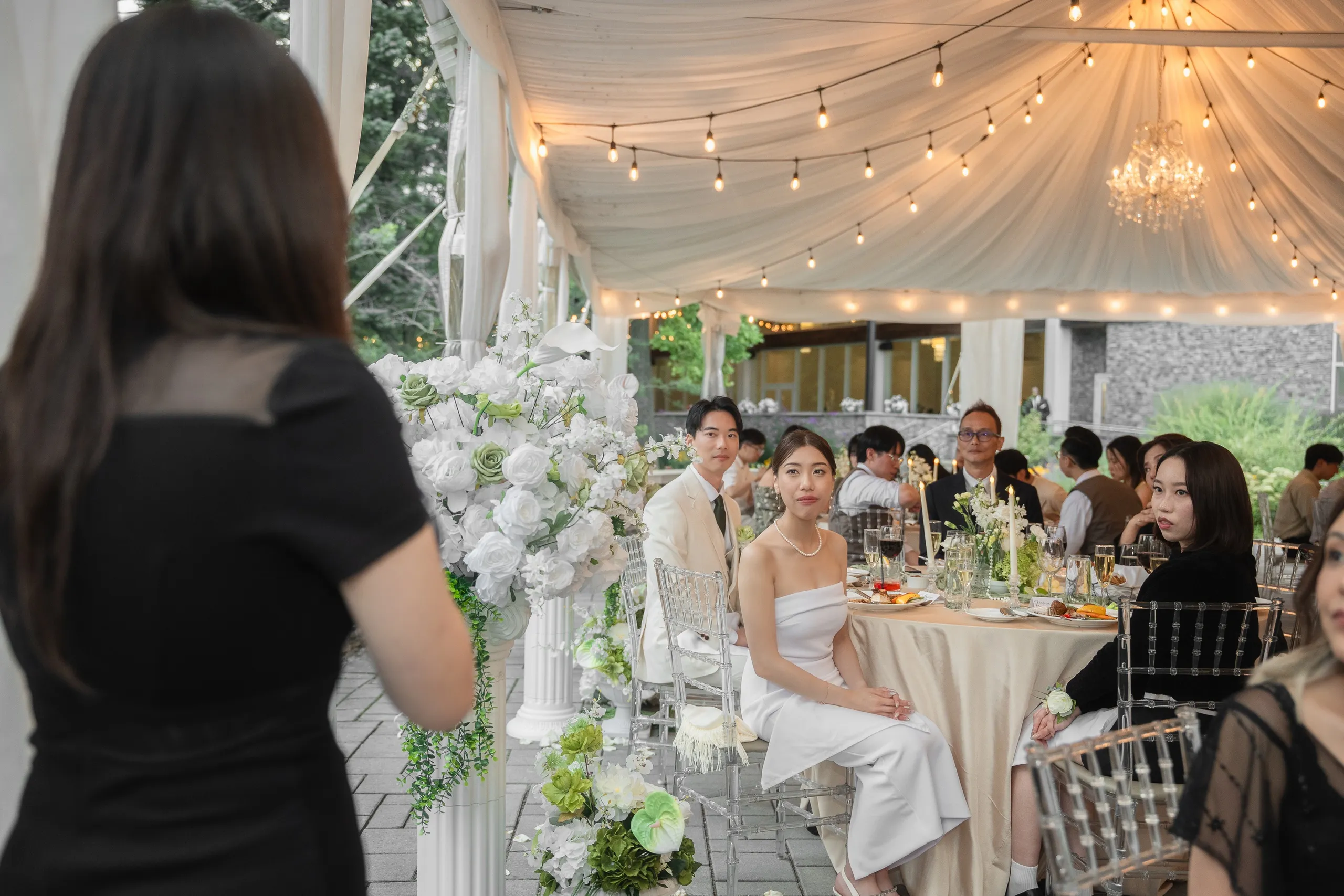 Framed past a guest's shoulder, Josie and Nelson seated at the head table under string lights, by AD Photography