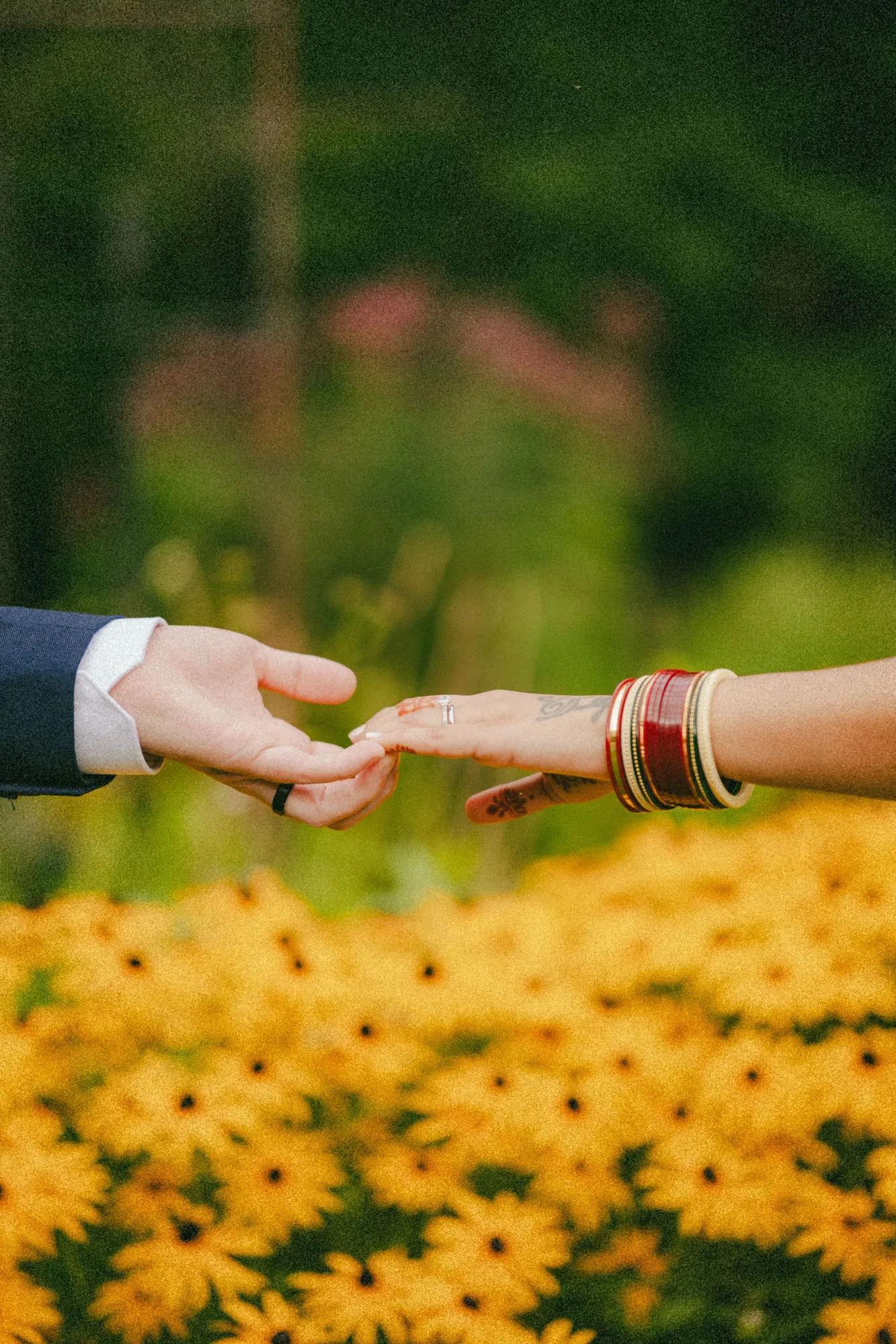 Fingertips about to touch over the yellow flower beds, her engagement ring catching the light, by AD Photography