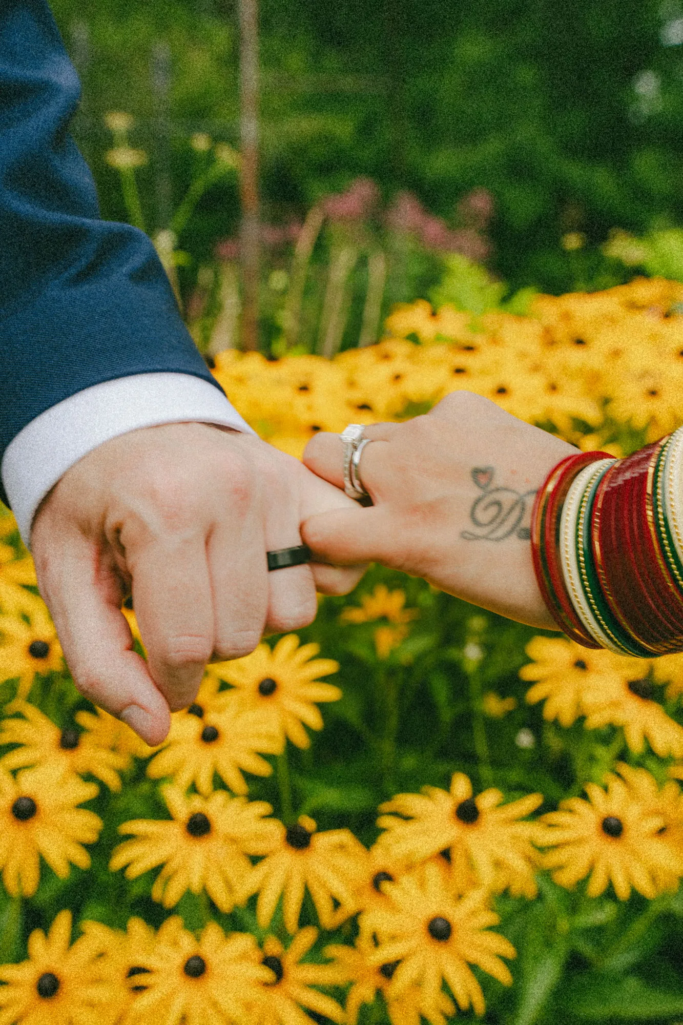 Grainy film style close-up of linked pinky fingers and rings above black eyed susans, by AD Photography