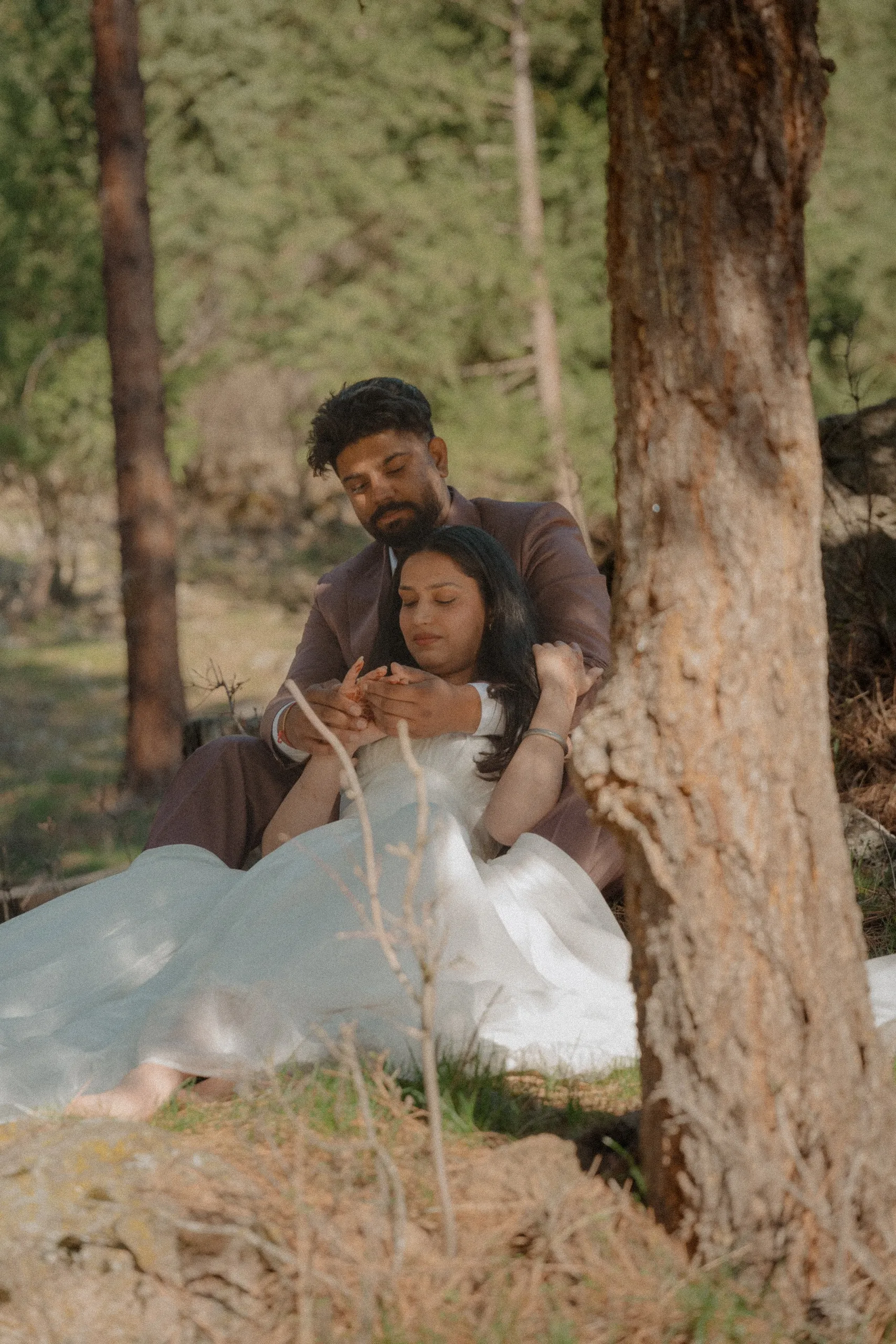 Framed past a pine trunk, Kamal holding the bride's hands with her eyes closed, by AD Photography