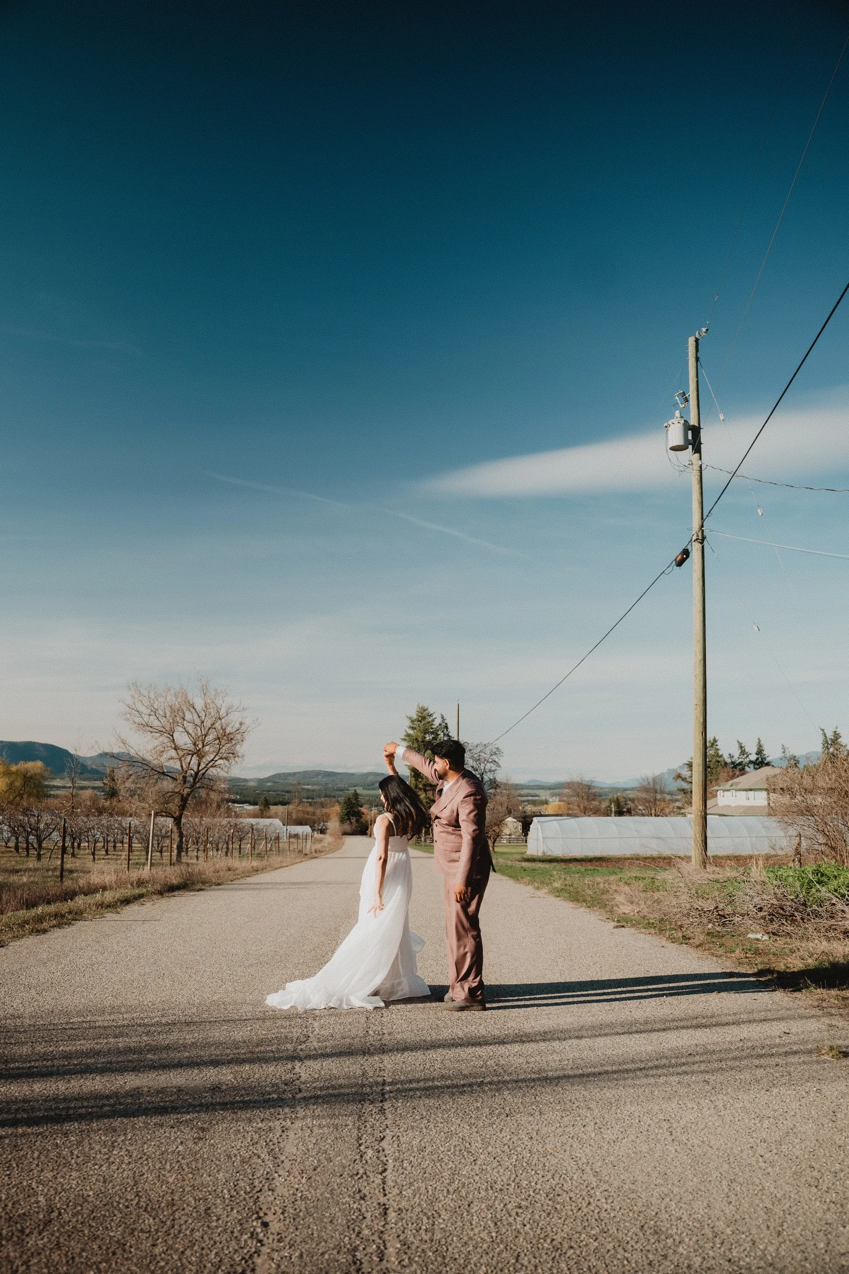 Kamal spinning Mehak mid road beneath a wide Okanagan sky and a lone power pole, by AD Photography