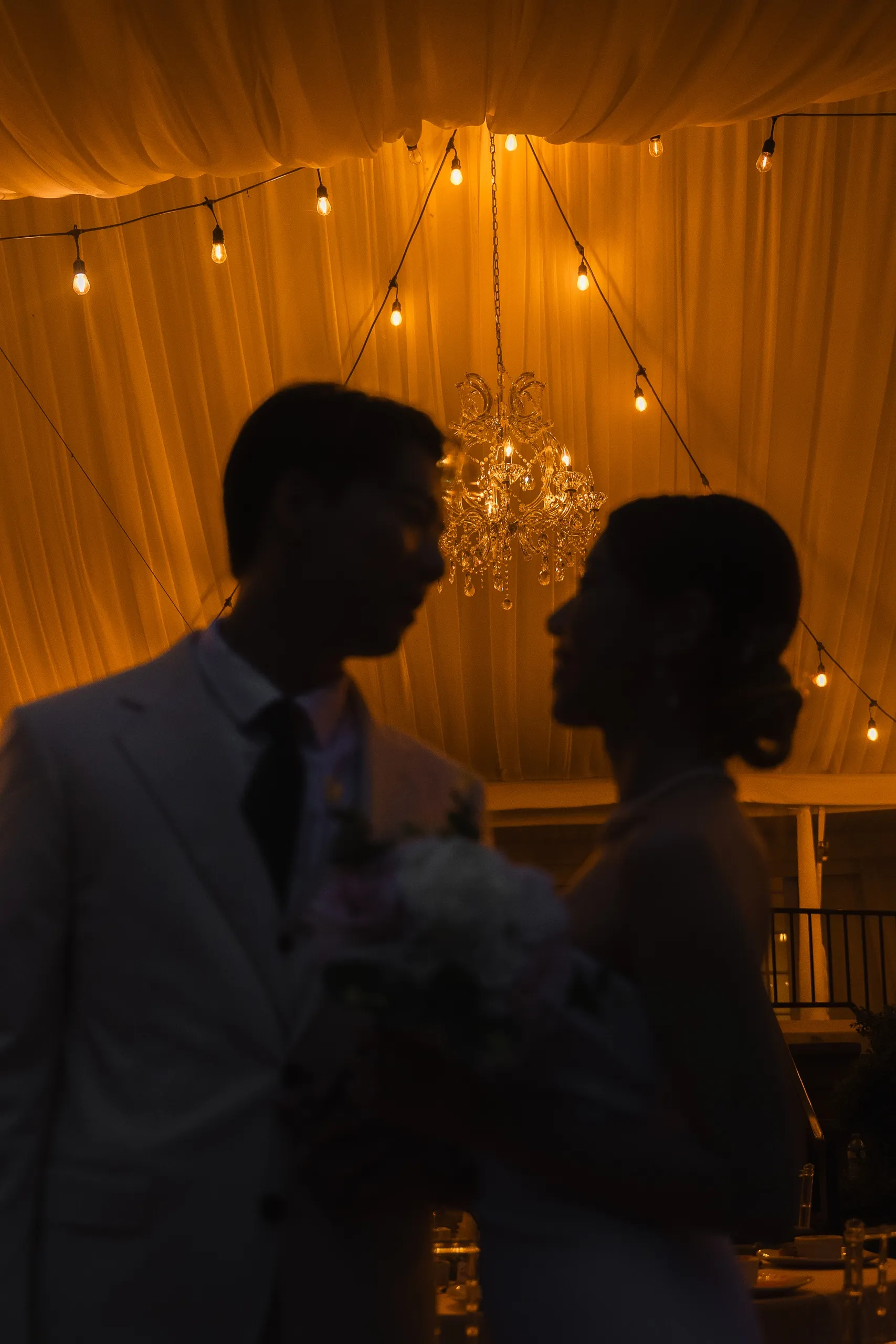 Silhouette of Josie and Nelson facing each other against the glowing chandelier and tent lights, by AD Photography