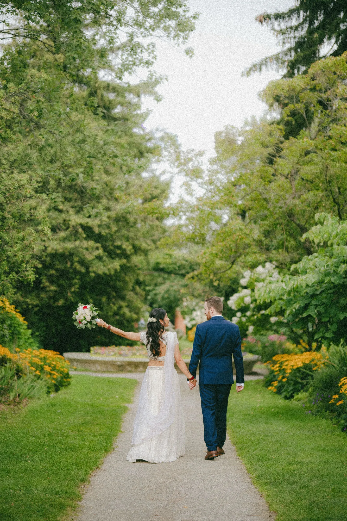 Nidhi raises her bouquet walking the flower lined path away from the camera with Derek, by AD Photography