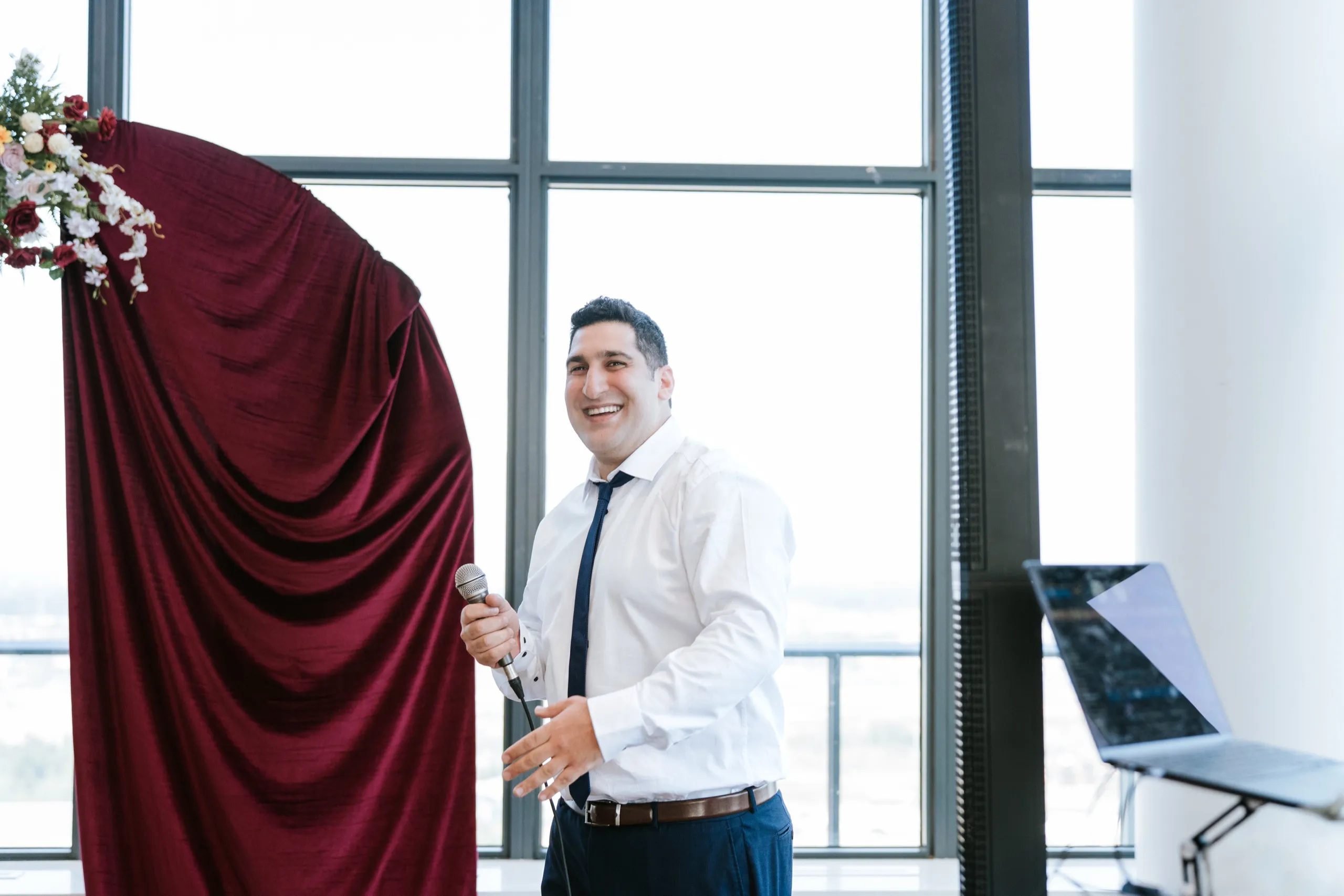 A family member takes the microphone in front of the burgundy drape and starts the speeches, by AD Photography