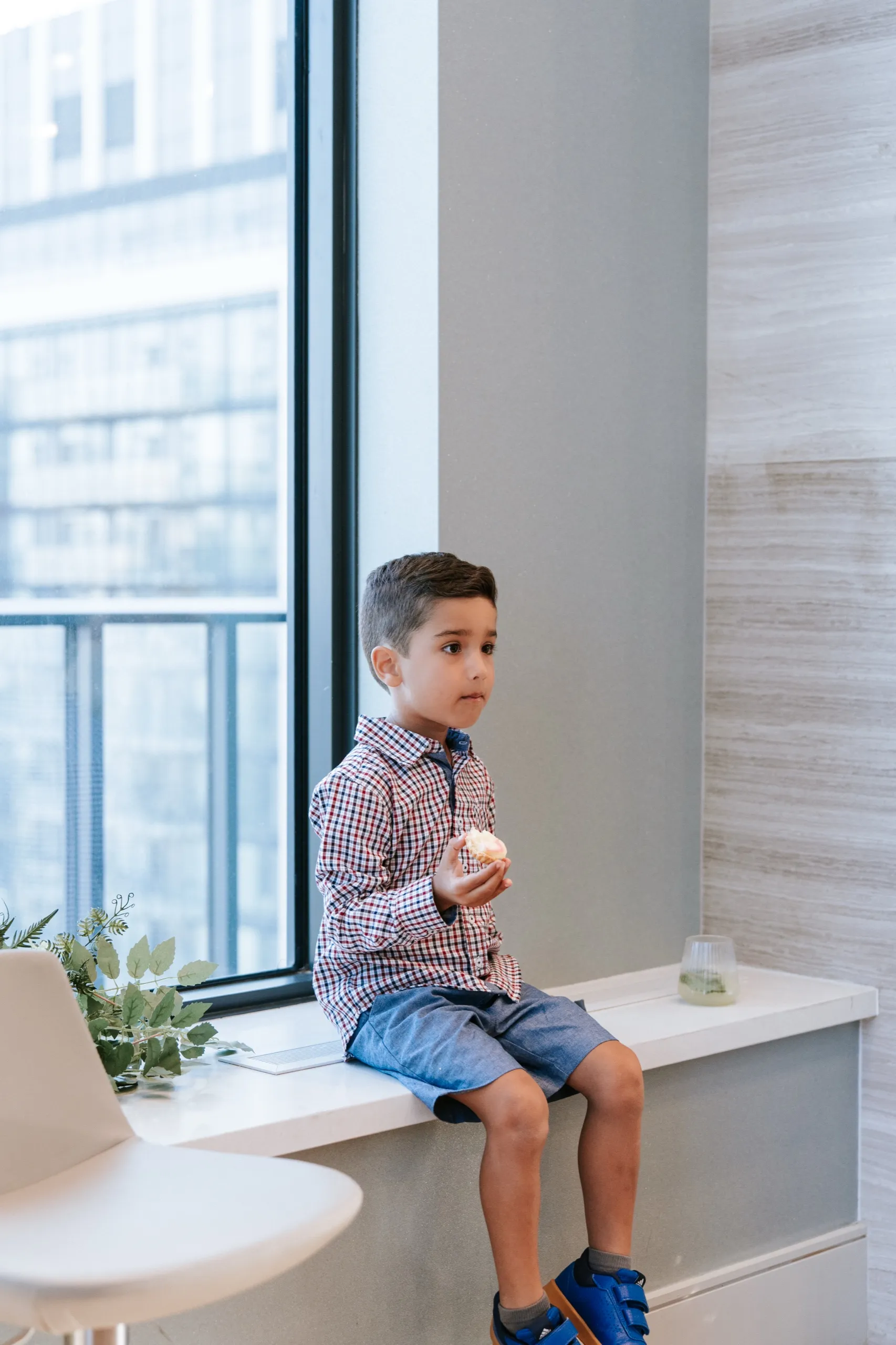 A small boy in a checked shirt sits on the window ledge working through a pastry, by AD Photography