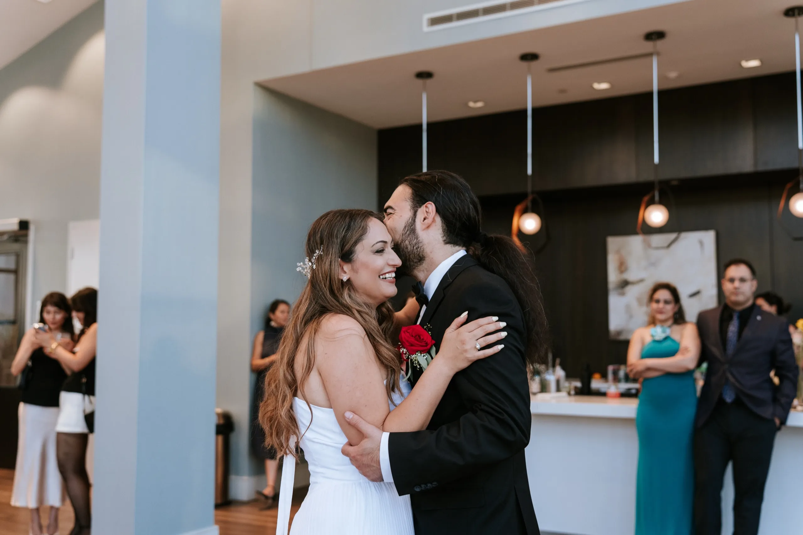 Moe kisses Zoha's cheek in front of the bar while guests look on, by AD Photography