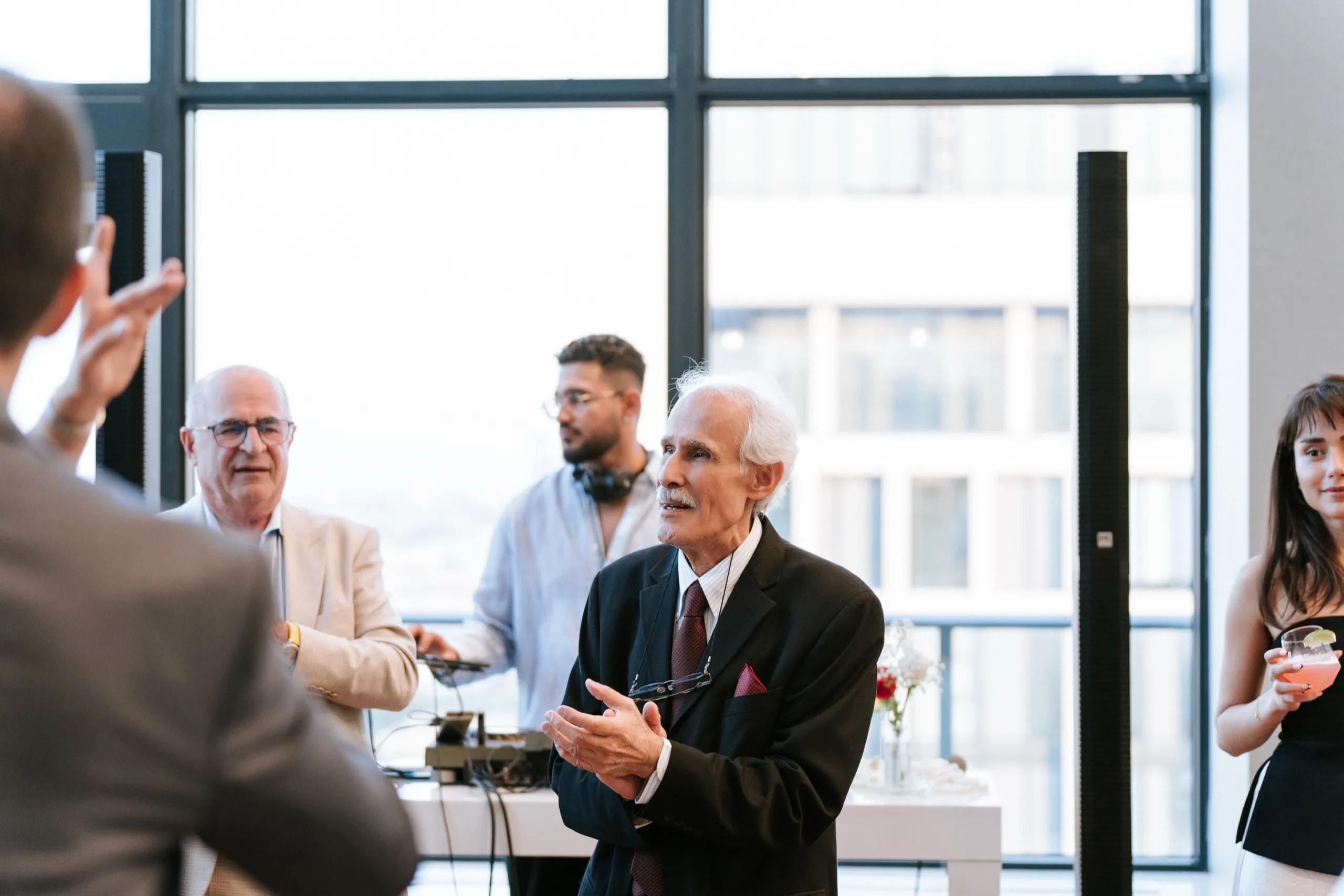 A family elder speaks to the room, guests listening against the wall of windows, by AD Photography