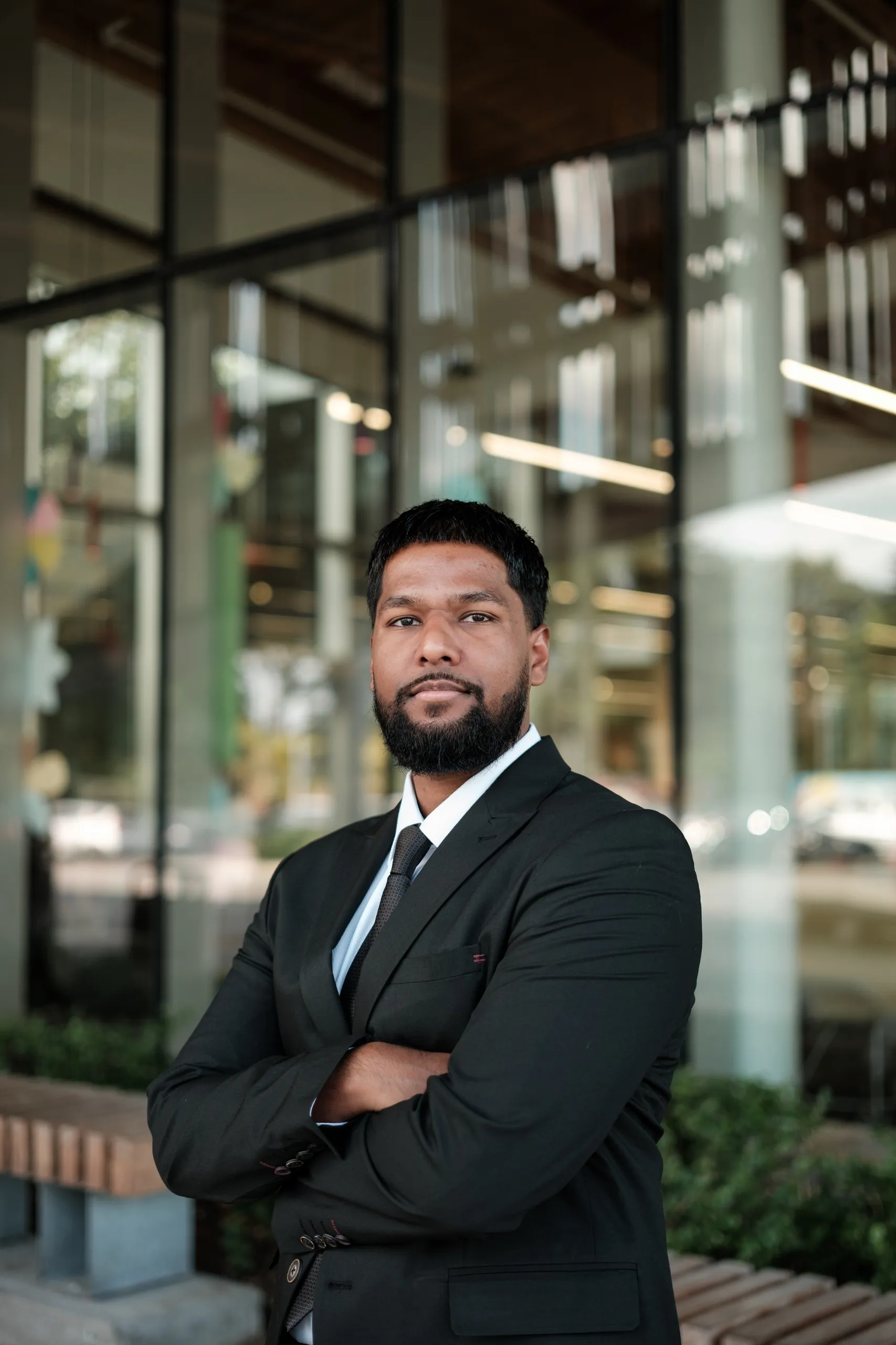 A headshot of a man with arms crossed and a composed expression outside Albion Public Library in Etobicoke, floor to ceiling glass behind him, by AD Photography