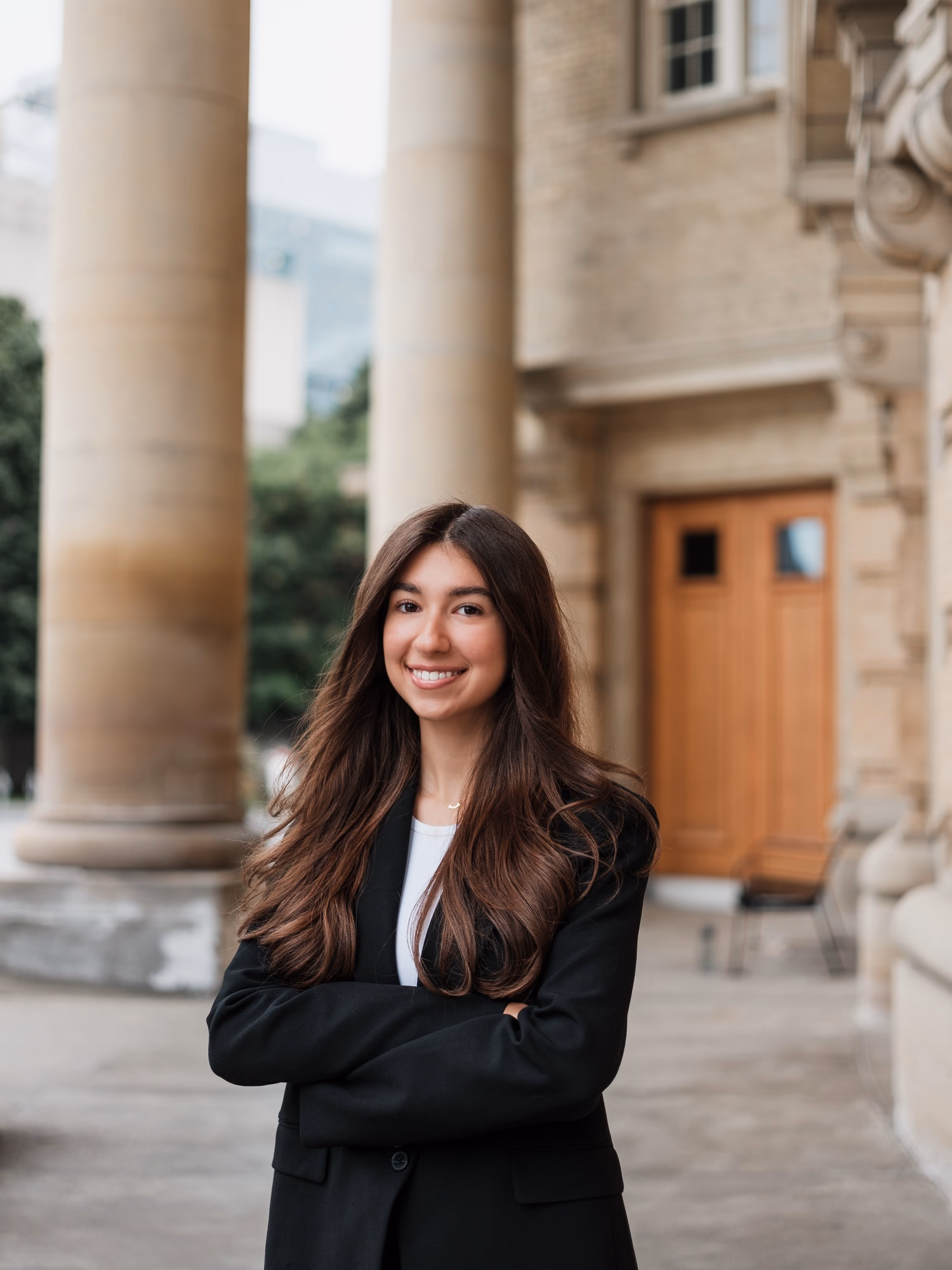 University of Toronto headshot of a young woman with long dark hair, smiling with her arms crossed in a black blazer and white top under the stone columns of the Convocation Hall portico, by AD Photography