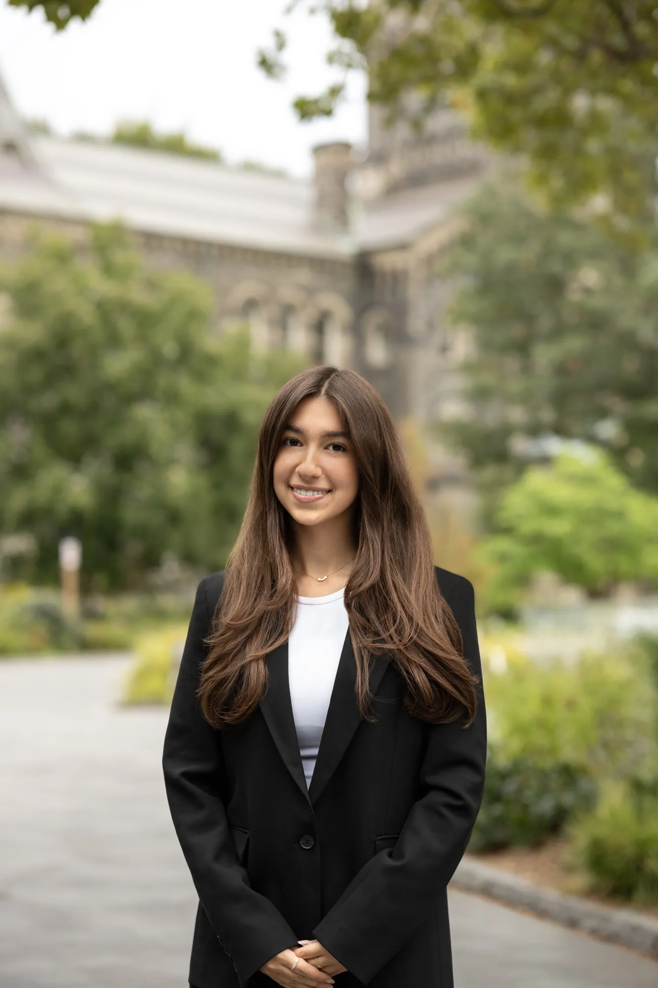 A young woman smiling with her hands clasped on a path outside University College at the University of Toronto, grey stone and green trees blurred behind her, by AD Photography