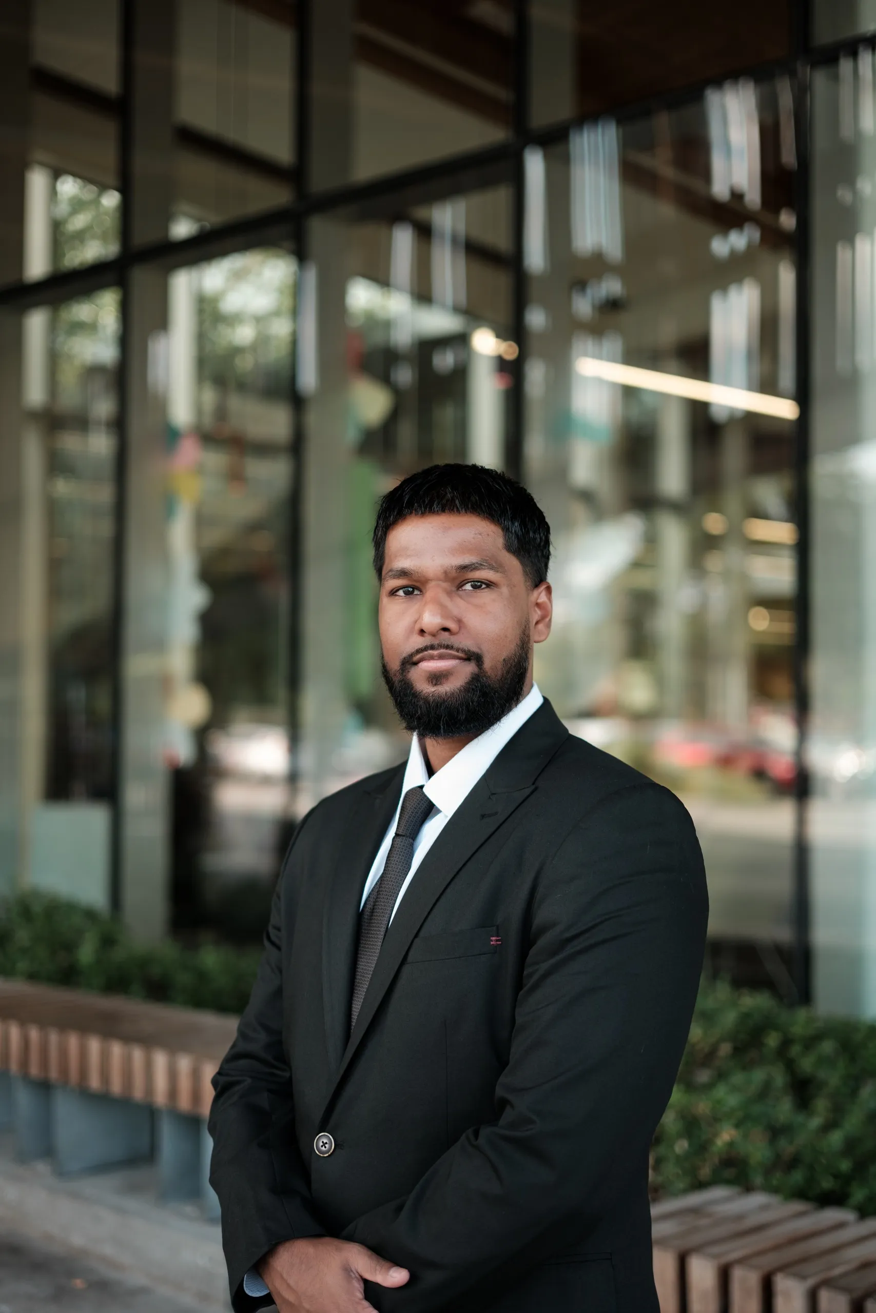 A man in a black suit standing with one hand in his pocket outside a modern Toronto building, a glass wall behind him, by AD Photography