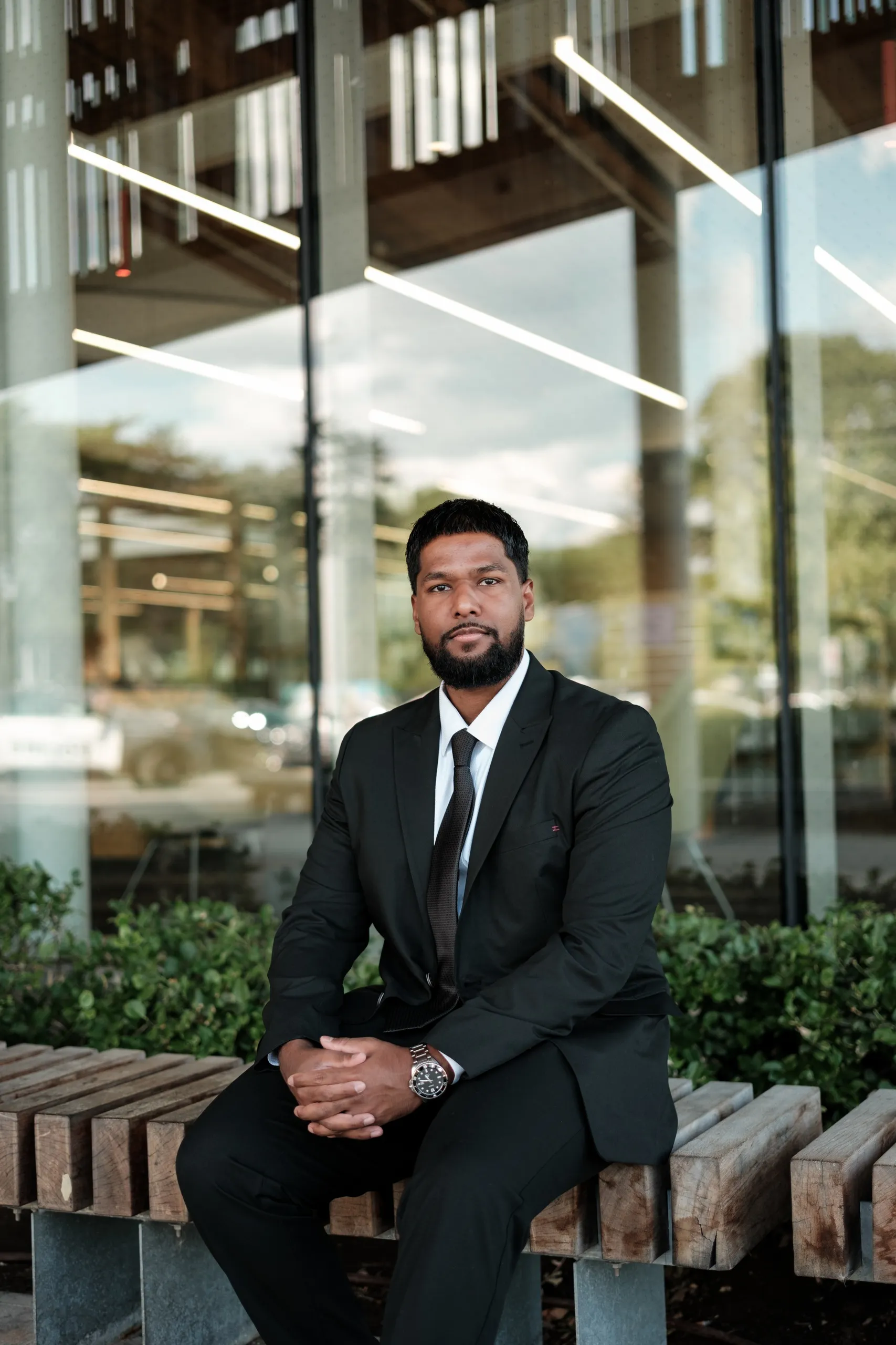 A man in a black suit seated on a wood bench outside a modern building in Toronto with a relaxed posture, by AD Photography