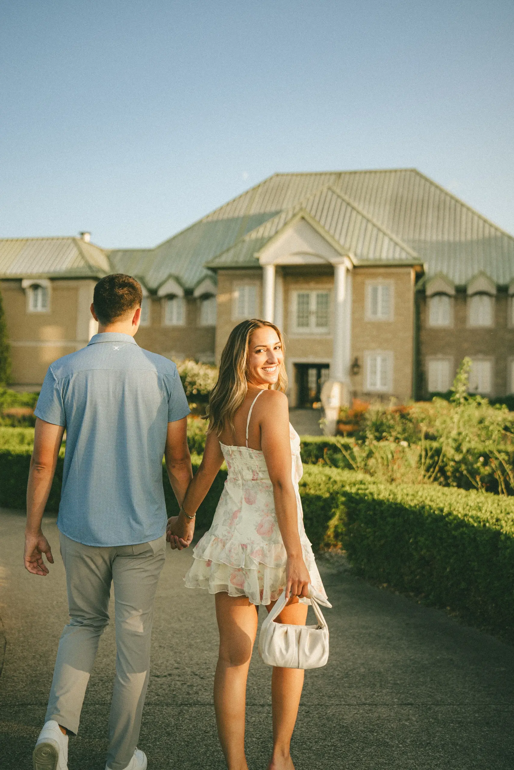 Pre-wedding couple walking hand in hand toward a stone manor at golden hour by AD Photography